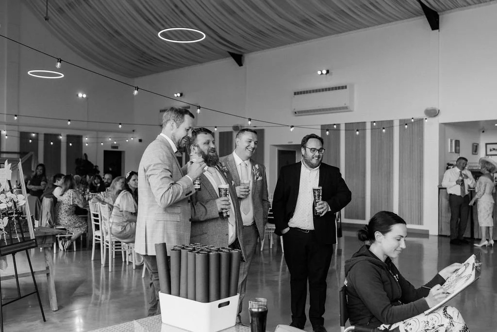 A group of five men in suits with drinks socializing at an event, while a woman sitting nearby draws guests in a decorated indoor venue.