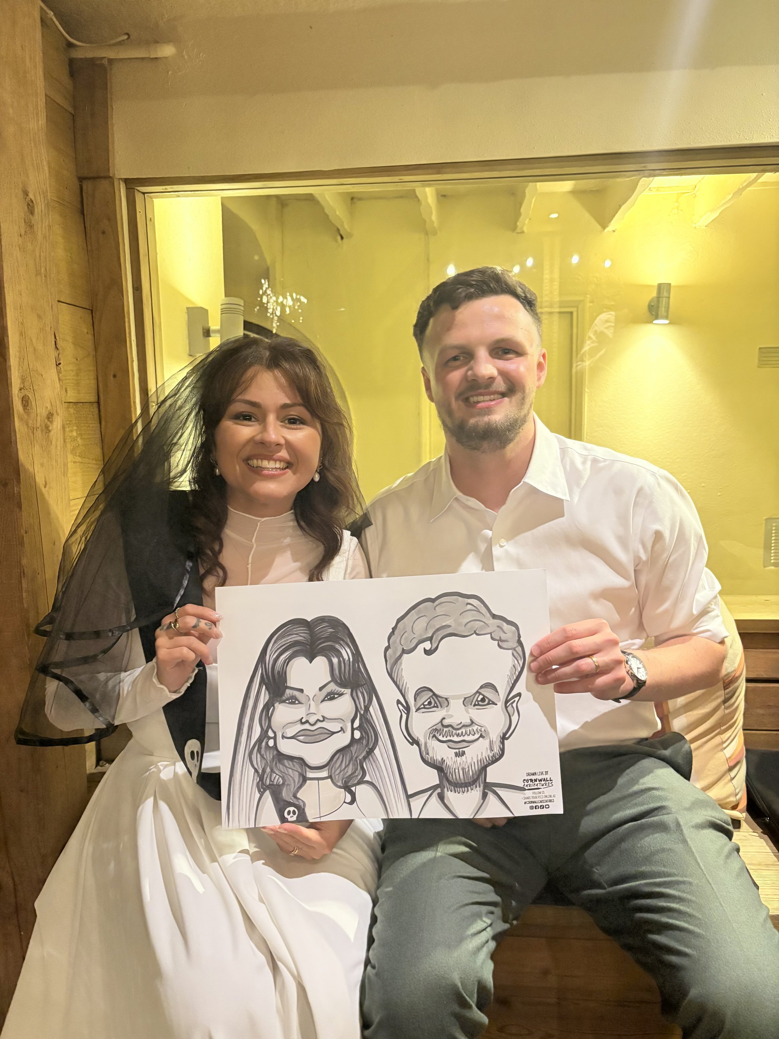 A smiling man and woman holding caricature drawings of themselves, seated indoors with yellow walls and wooden accents.