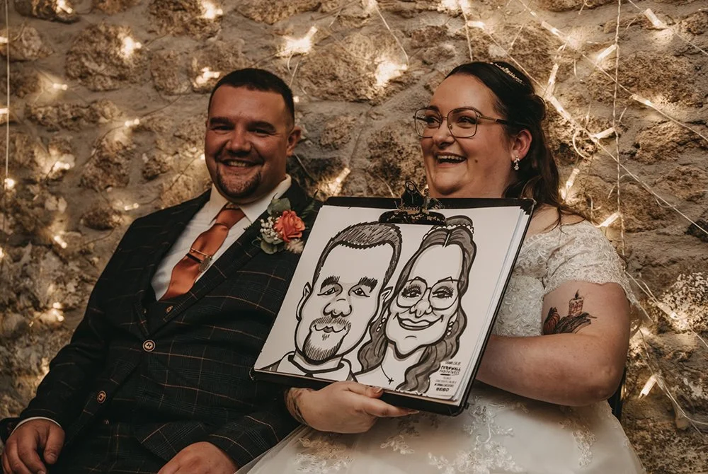 A bride and groom smiling, holding a caricature drawing of themselves at their wedding reception with a stone wall and fairy lights in the background.