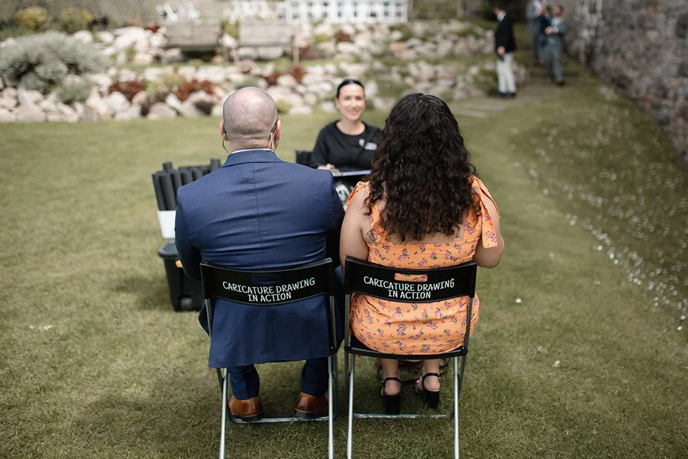 Two people seated outdoors at a table with a woman drawing caricatures of them, on chairs labeled "Caricature Drawing In Action," while two others stand nearby in the background near a stone wall and water feature.