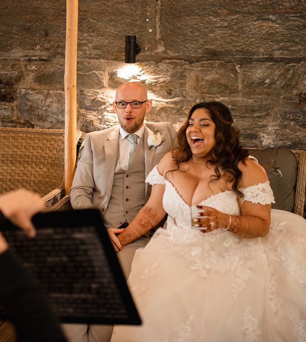 Bride and groom sitting on a couch during their wedding reception, laughing and smiling, with a rustic stone wall in the background.