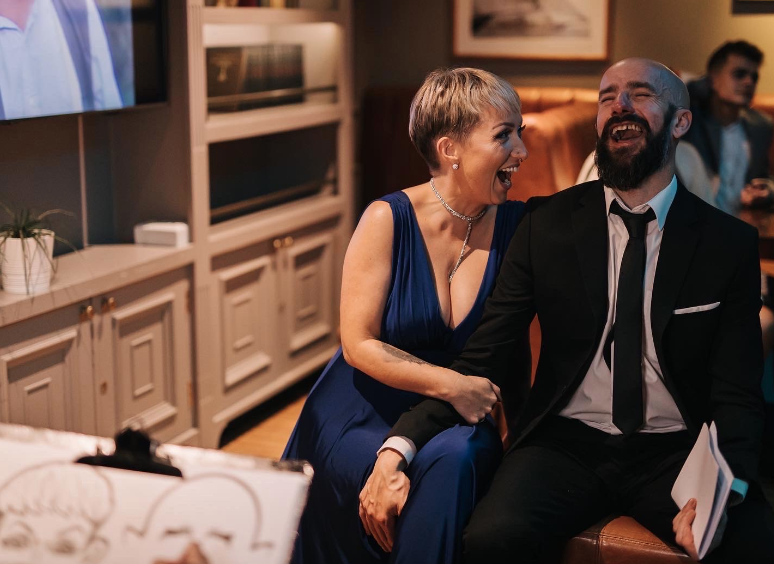 A woman in a deep blue dress and a man in a black suit with a tie are sitting closely, smiling and sharing a joyful moment at a social event in a warmly lit room.