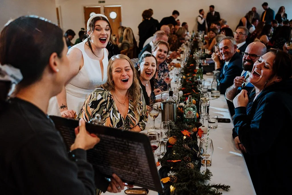 People at a long banquet table laughing and enjoying a wedding reception, with a woman showcasing a caricature drawn live.