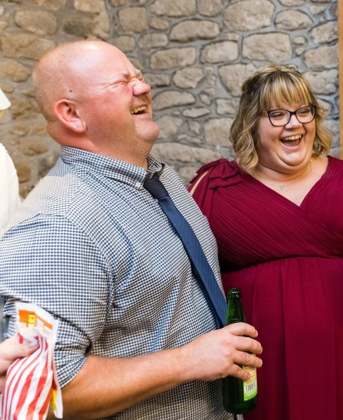 Two adults laughing joyfully at an indoor gathering, one holding a beer bottle, with a stone wall in the background.