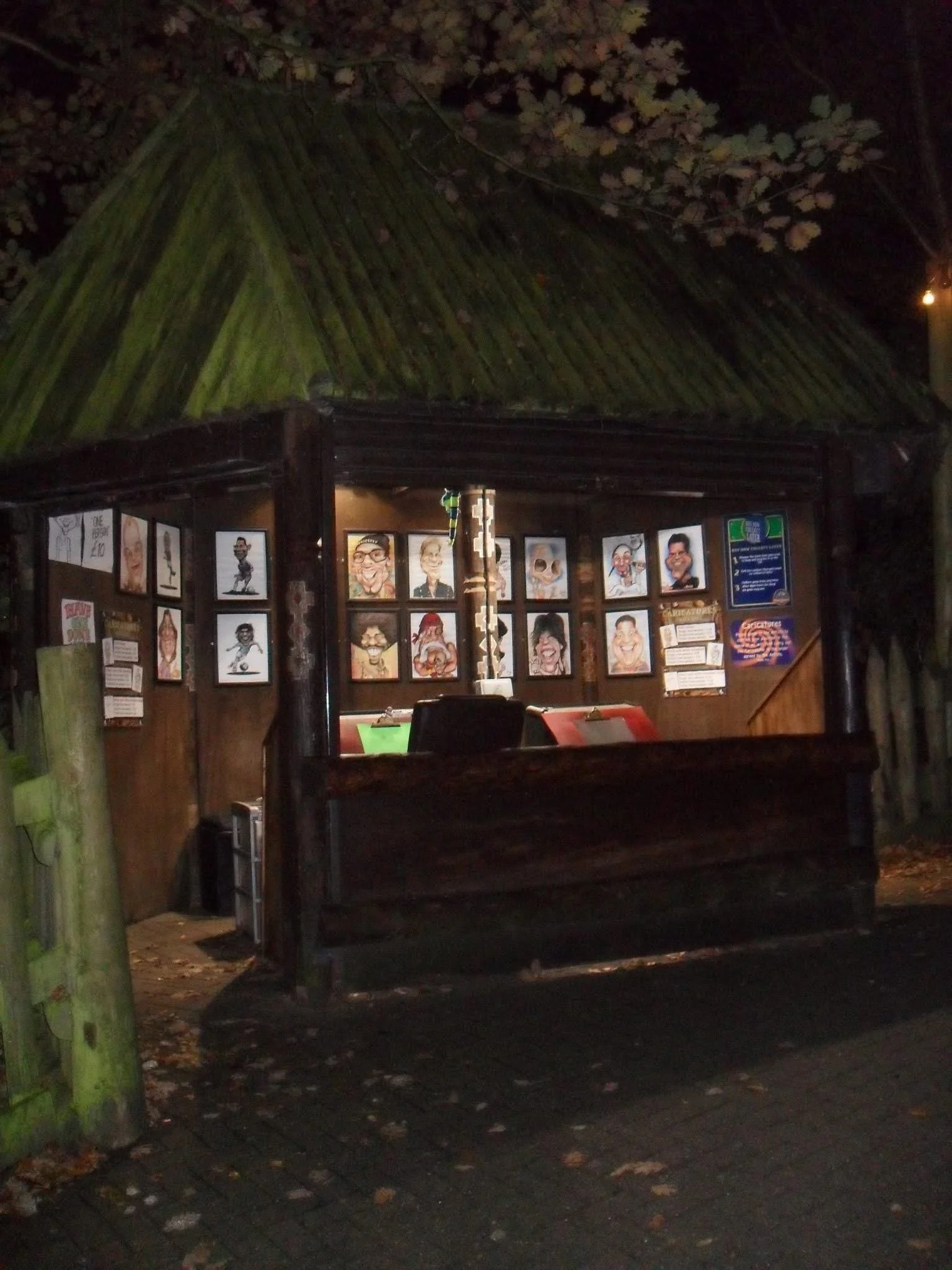 Nighttime scene of a small wooden pavilion or shelter with caricature portraits on the walls, surrounded by trees with fallen leaves on the ground.