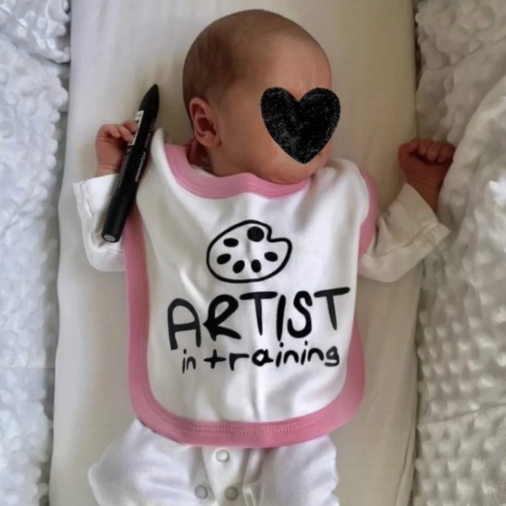 A baby lying on a blanket, wearing a white bib with pink trim that says 'Artist in Training' and holding a black marker. The baby's face is covered with a black heart sticker.
