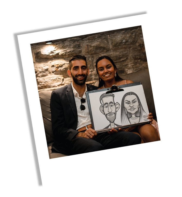 A smiling couple sitting on a sofa, holding a drawing of themselves with exaggerated caricature features, in a cozy indoor setting with a stone wall background.