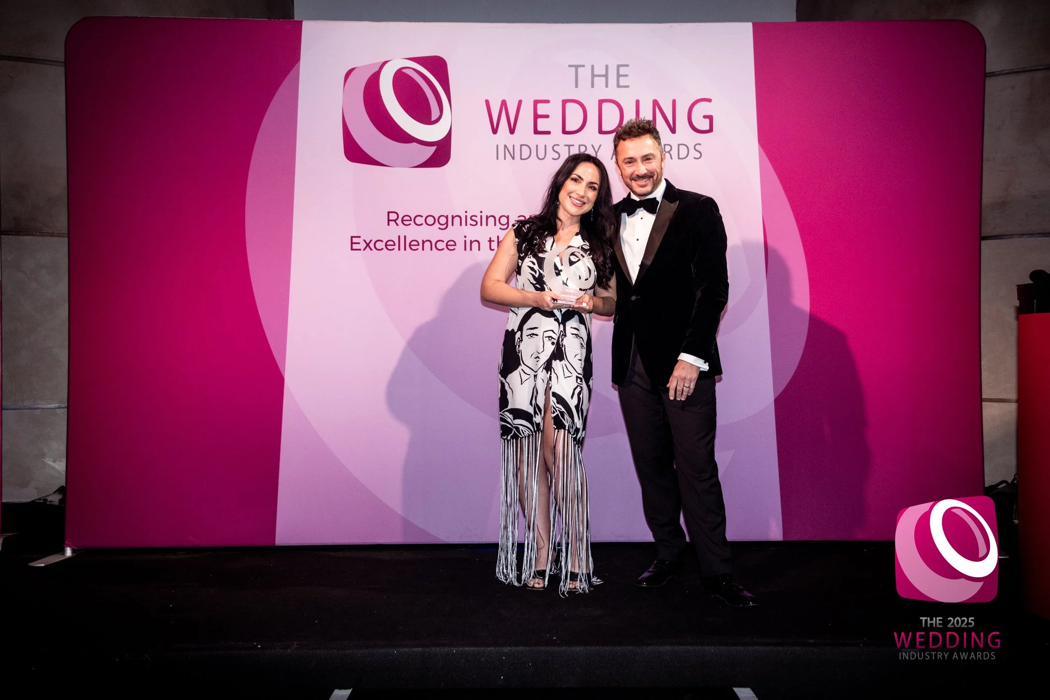 A man and a woman standing together on a stage with a pink and white backdrop at the Wedding Industry Awards. The woman is holding a trophy and wearing a black-and-white dress with a graphic face print. The man is dressed in a black tuxedo with a bow tie, smiling at the camera.