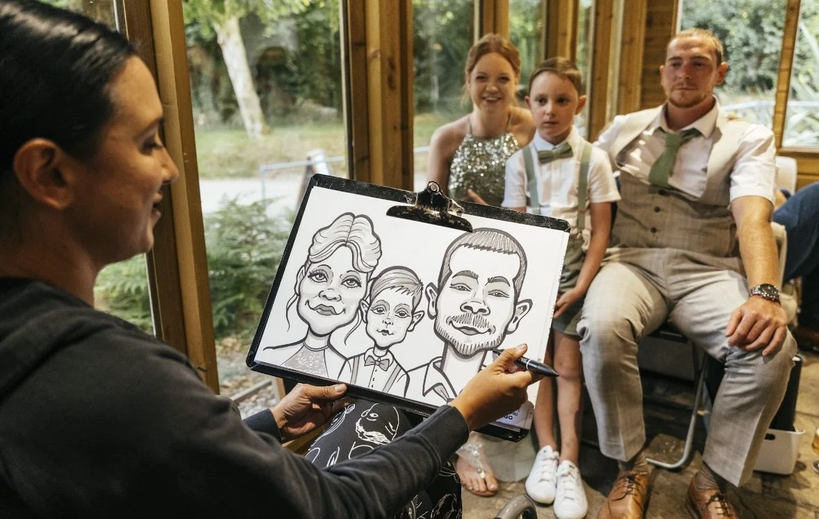 A caricature artist drawing a cartoon of a family with a woman, one man, and a young boy, during a gathering in a room with large windows showing outdoor greenery.