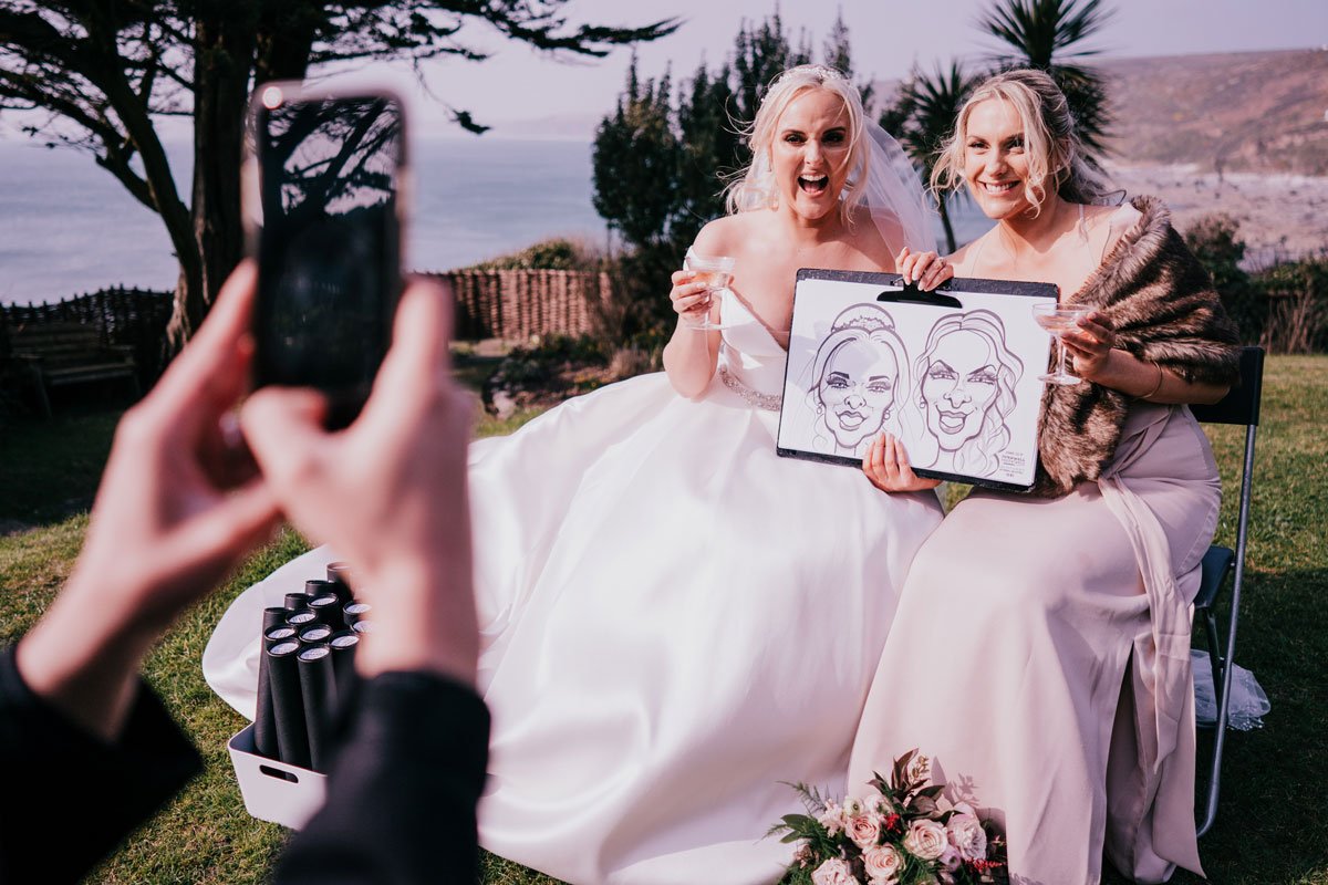 Two women in wedding dresses are sitting outdoors near water, holding glasses and a sketch of their faces, with one woman showing an excited expression and the other smiling, while someone takes their photo.