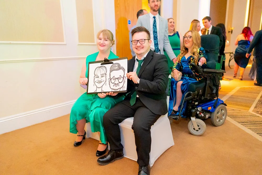 Group of people at a wedding or formal event, with one person in a wheelchair. The main couple is holding a caricature portrait. There are smiling women and men in the background.