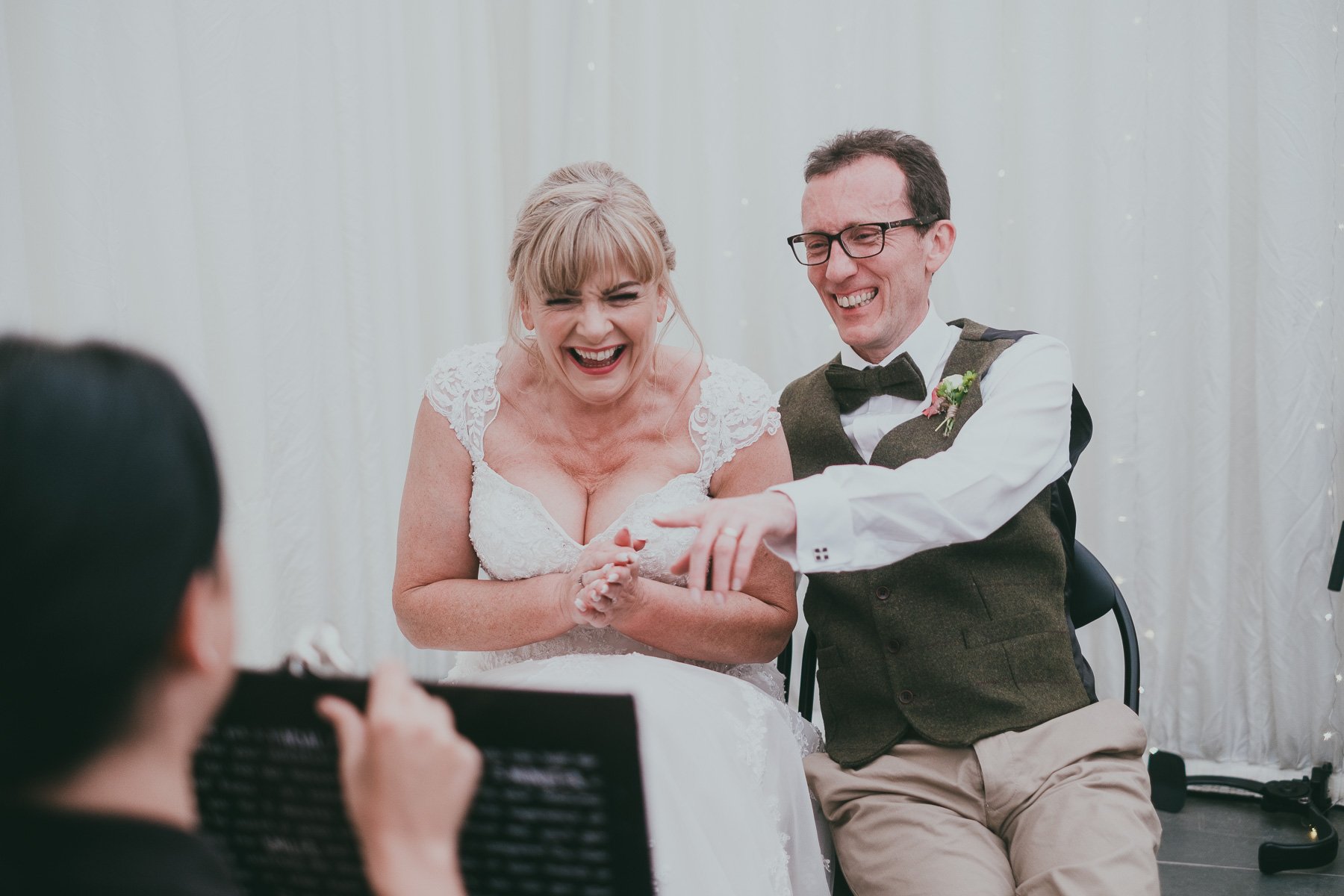 A joyful bride in a white lace wedding dress and a groom in a vest and bow tie smiling and laughing at their caricature during a wedding celebration.