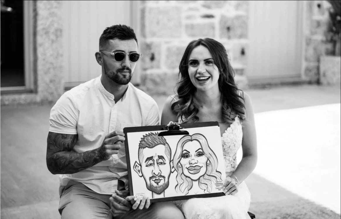 A man with sunglasses and a woman in a white dress sitting outdoors, holding a caricature portrait of a man and woman. The background features a stone wall and a door.