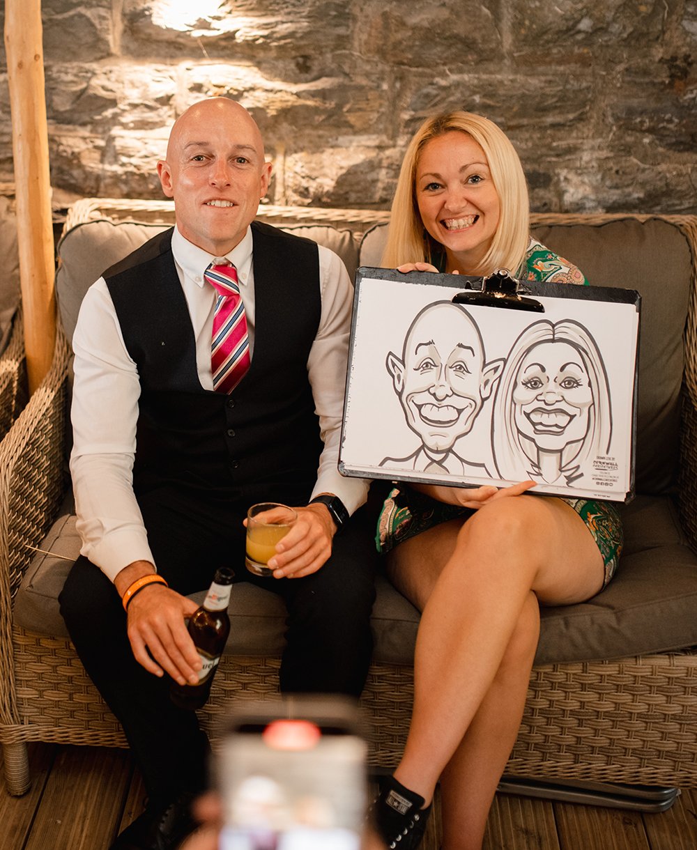 A man and woman sit on a wicker couch, smiling and holding a caricature drawing of themselves. The man is bald, dressed in a vest and tie, holding a drink and a beer. The woman has blonde hair, wearing a colorful dress, with her legs crossed, smiling