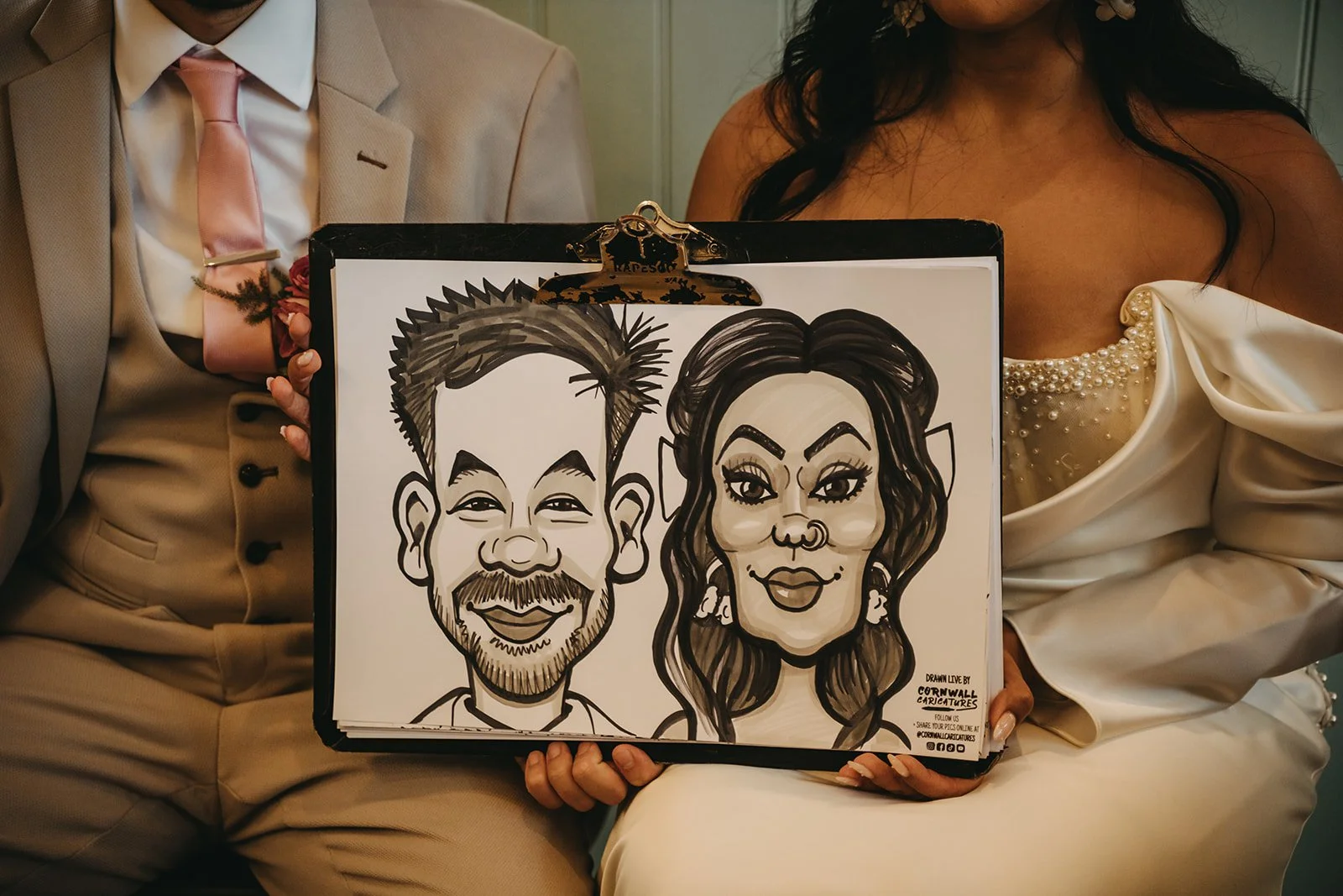 A person in a suit and a woman in a dress are sitting close together, holding a portrait drawing of them as cartoon characters.