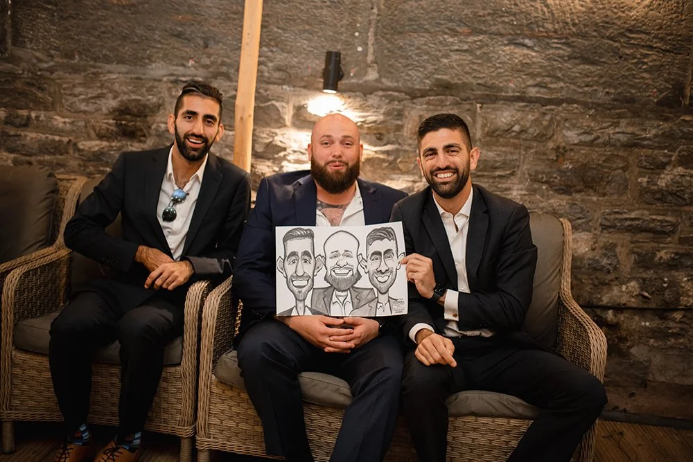 Three men in suits sitting on a wicker sofa in front of a stone wall, with one holding a caricature drawing of the three of them.