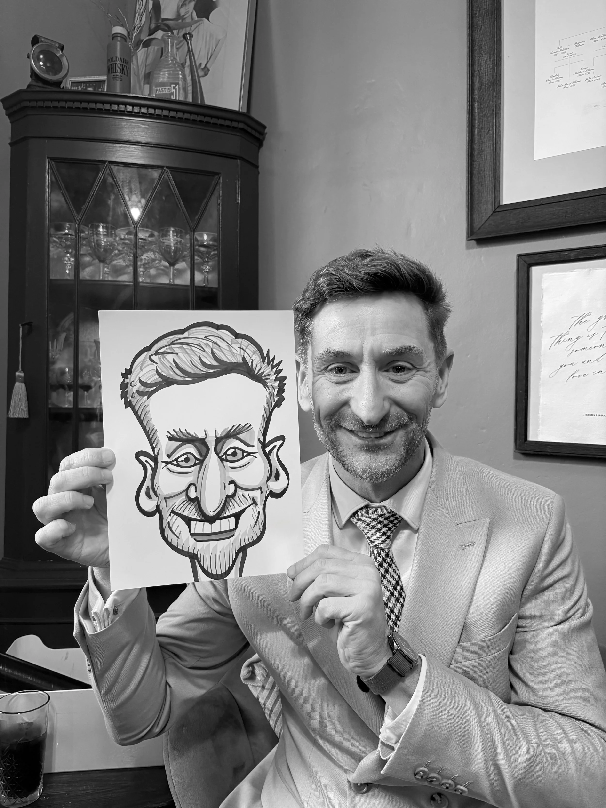 A man in a suit and tie is sitting at a table, holding a caricature drawing of himself that depicts him with exaggerated facial features and hair. The man is smiling and looking at the camera. There is a glass of dark beverage on the table beside him.