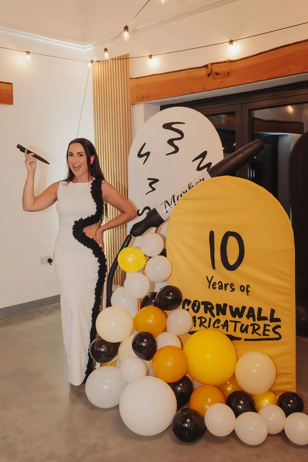 A woman in a white dress with black ruffle accents standing next to a sign celebrating 10 years of Cornwall Caricatures, with balloons and a large fake ink pen.