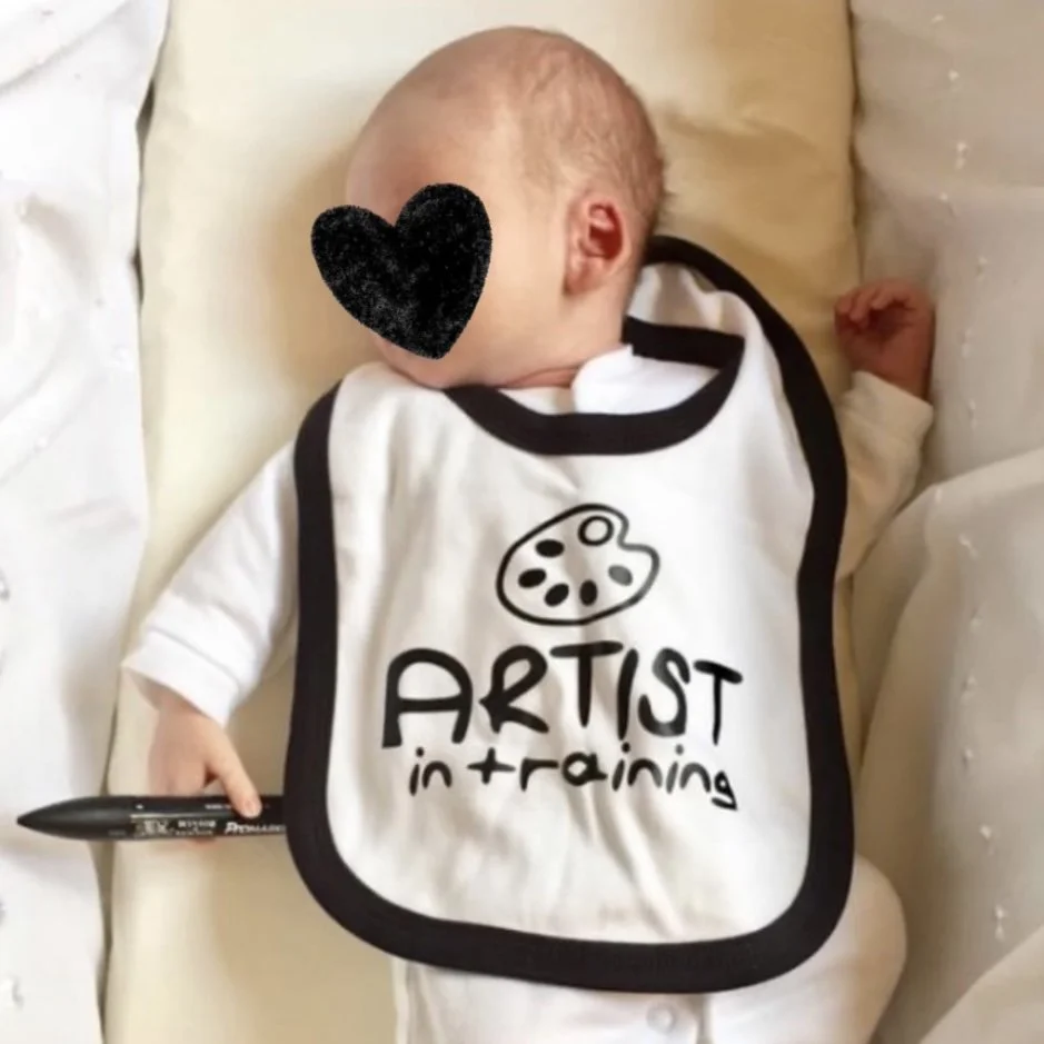A baby with a black heart covering its face, wearing a bib that says 'ARTIST in training' with a paint palette graphic, lying on a bed with a black marker in hand.