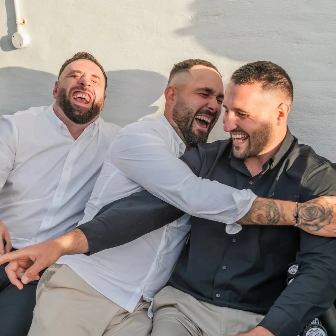 Three men laughing and hugging each other as they see their live caricature reveal, sitting outdoors against a light-coloured wall, enjoying a joyful moment at a wedding.