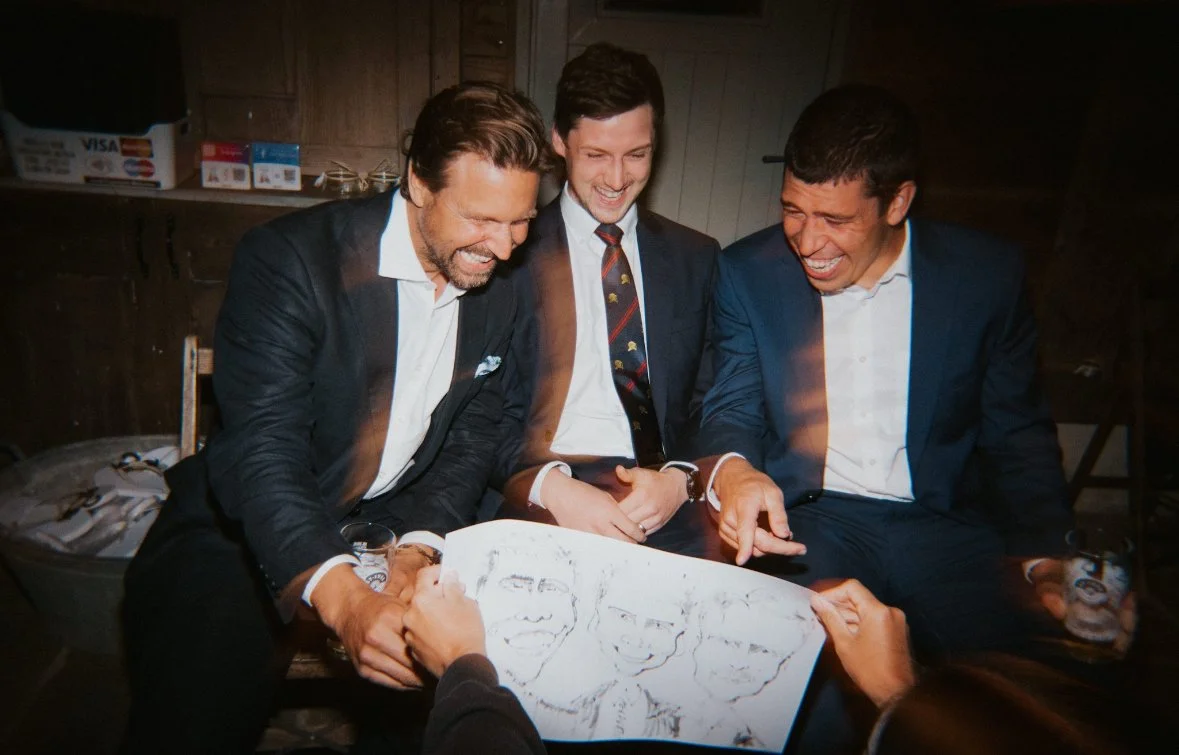 Three men in suits laughing and looking at a drawing of their faces being held by someone off-camera.