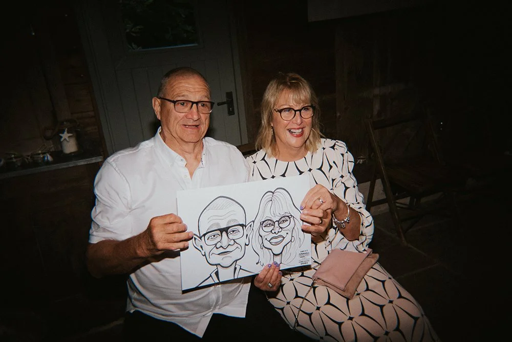A man and woman sitting together indoors, smiling and holding a caricature drawing of themselves, both wearing glasses and dressed in light-colored clothing.