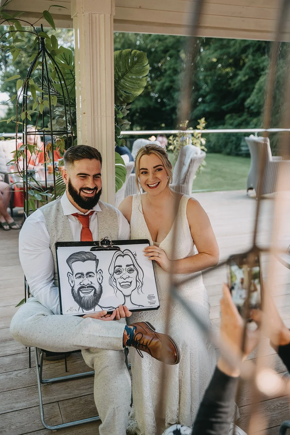 A smiling couple, a man with a beard and a woman in a wedding dress, holding a caricature drawing of themselves, at an outdoor event or wedding reception.