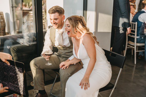 A bride and groom sitting closely together, smiling and laughing at a wedding celebration, with a guest holding a caricature up in front of them.