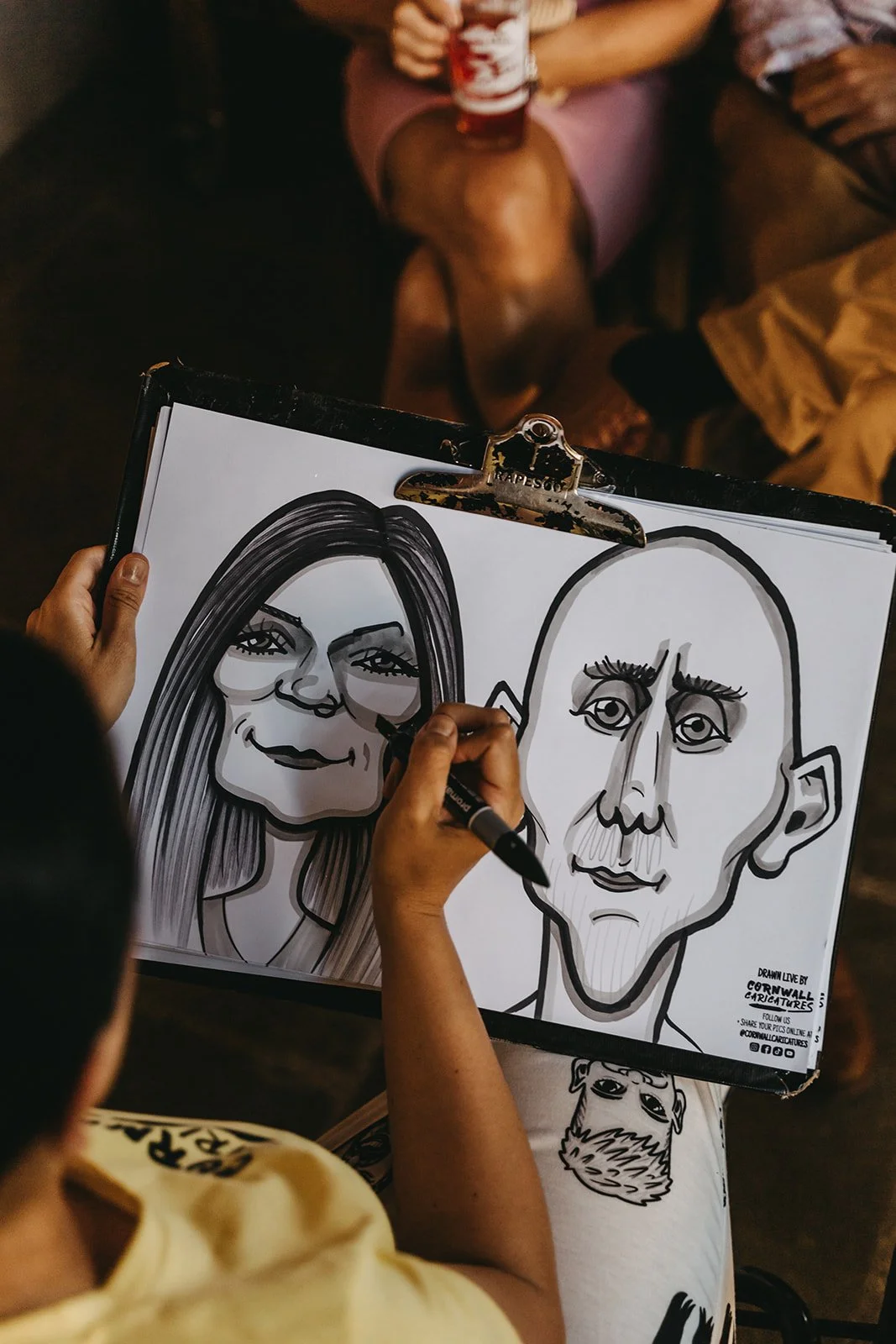 Person drawing a caricature of a woman and a man on a sketchpad. The woman has long hair and glasses, and the man is bald.