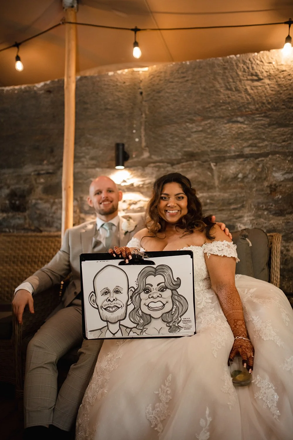 A newlywed couple sitting together, the bride holding a caricature drawing of themselves, smiling at the camera inside a rustic venue with string lights.