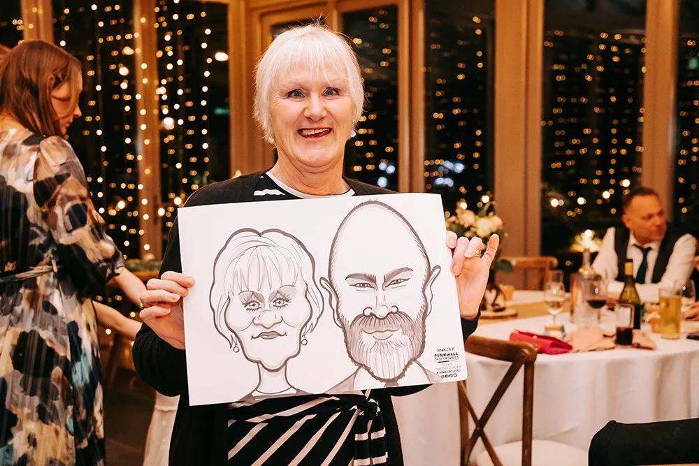 An older woman smiling and holding up a caricature drawing of herself and a man. She is at a decorated indoor event with string lights and table settings.