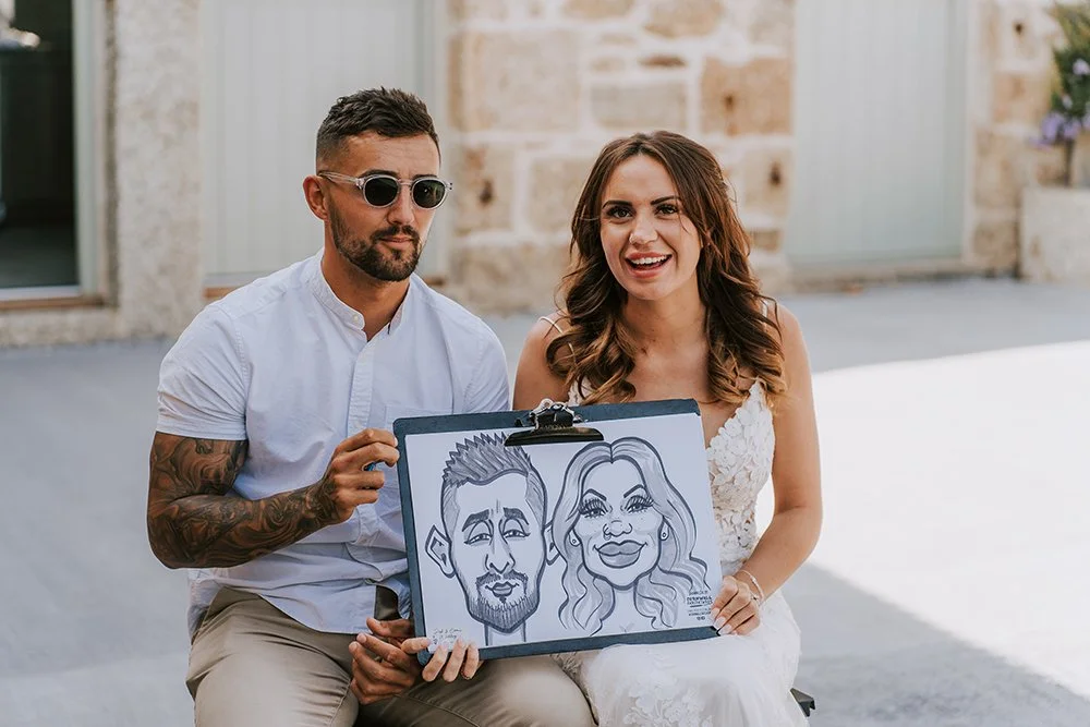 A smiling woman in a white dress sitting next to a man with sunglasses, holding a caricature of themselves, outdoors in front of a brick building.