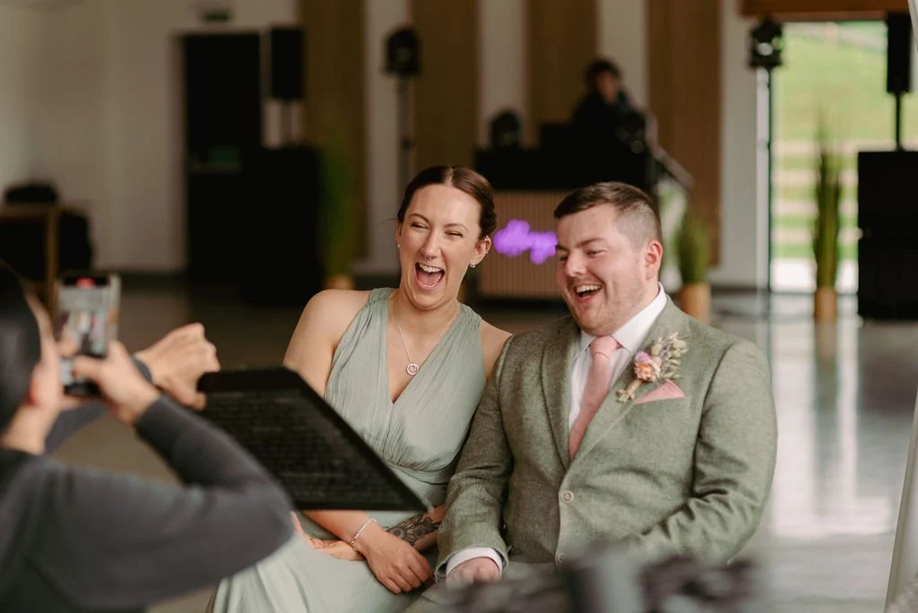 A woman and a man laughing and sitting close together during a wedding or special event, with a person taking a photo in the foreground as they see their caricature.