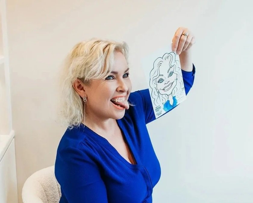 A woman with blonde hair, wearing a blue top, holding a caricature portrait of herself, sticking out her tongue, and smiling.
