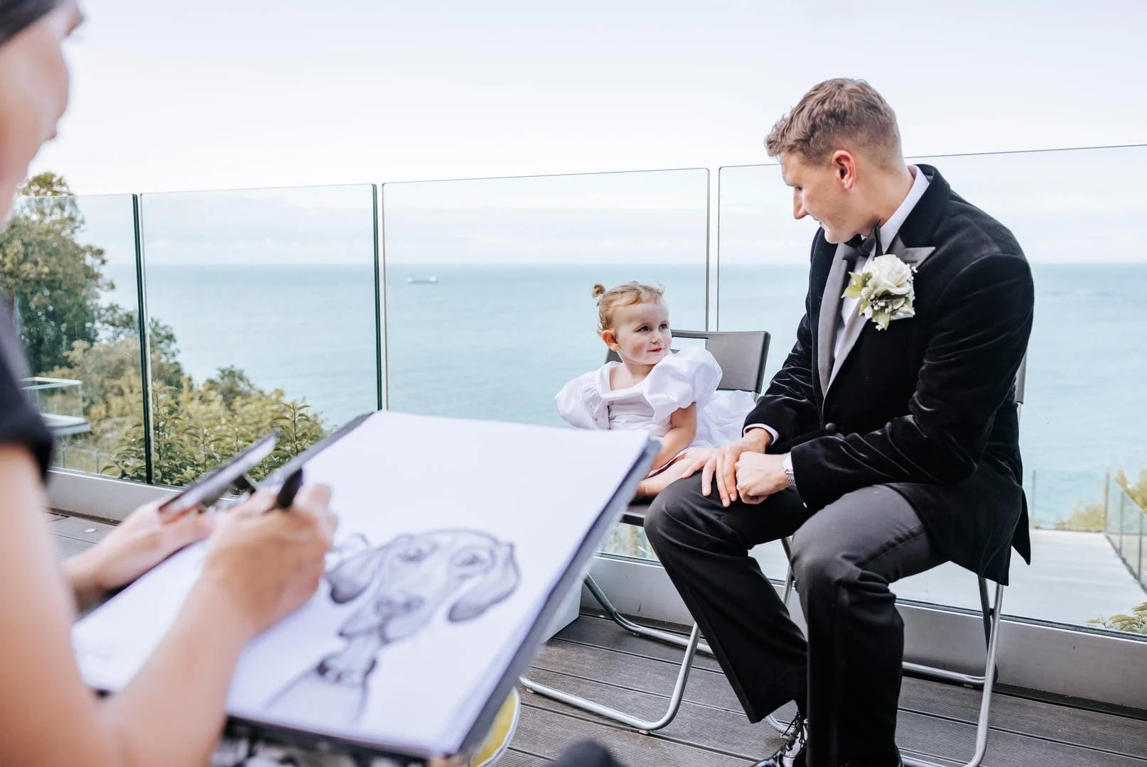 A young girl in a white dress being drawn by an artist, with a man in a tuxedo sitting next to her on a balcony overlooking the ocean.