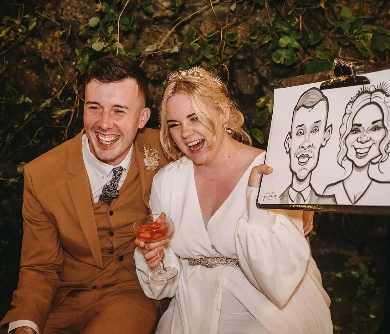 A smiling man and woman sitting together, the woman is holding a glass of drinks and a caricature drawing of themselves. They are dressed in formal or wedding attire, with the woman in a white dress and the man in a brown suit. The woman is wearing a headband or tiara, and the man has short dark hair. The background consists of greenery and rocks.
