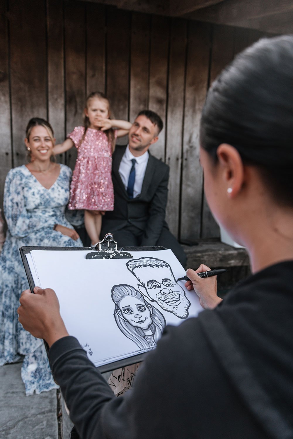 Cartoonist drawing caricatures of a family of three, with a woman in a blue dress and a man in a suit, and a little girl in a pink dress, during a outdoor event with a wooden background.