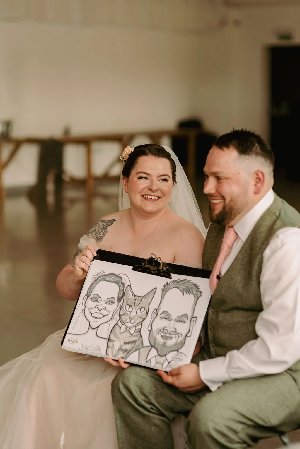 A bride and groom sitting together, smiling, with the bride holding a framed caricature of herself, a cat, and the groom.