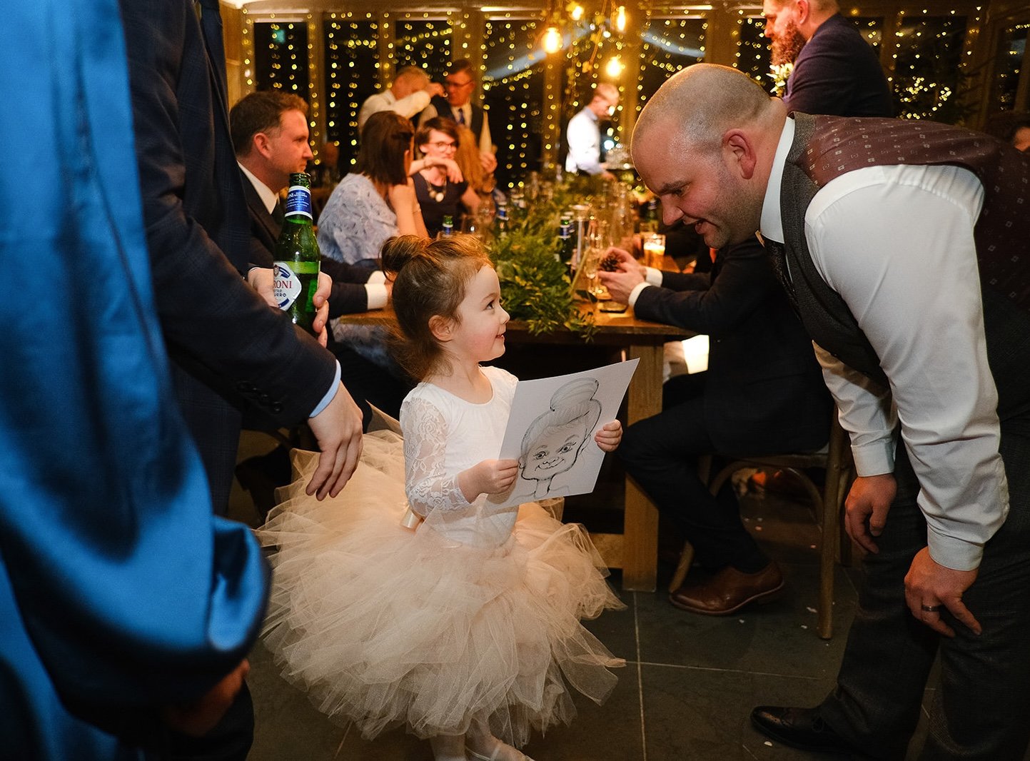 A young girl in a white lace dress and a tulle skirt is smiling at a man leaning towards her, who is dressed in a white shirt and gray vest. The girl is holding a piece of paper with a drawing of a smiling face. They are at a festive gathering with p