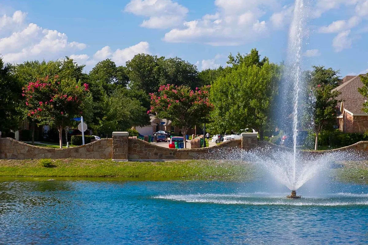 A park pond with a fountain in the center, surrounded by trees, houses, and a stone wall in the background.
