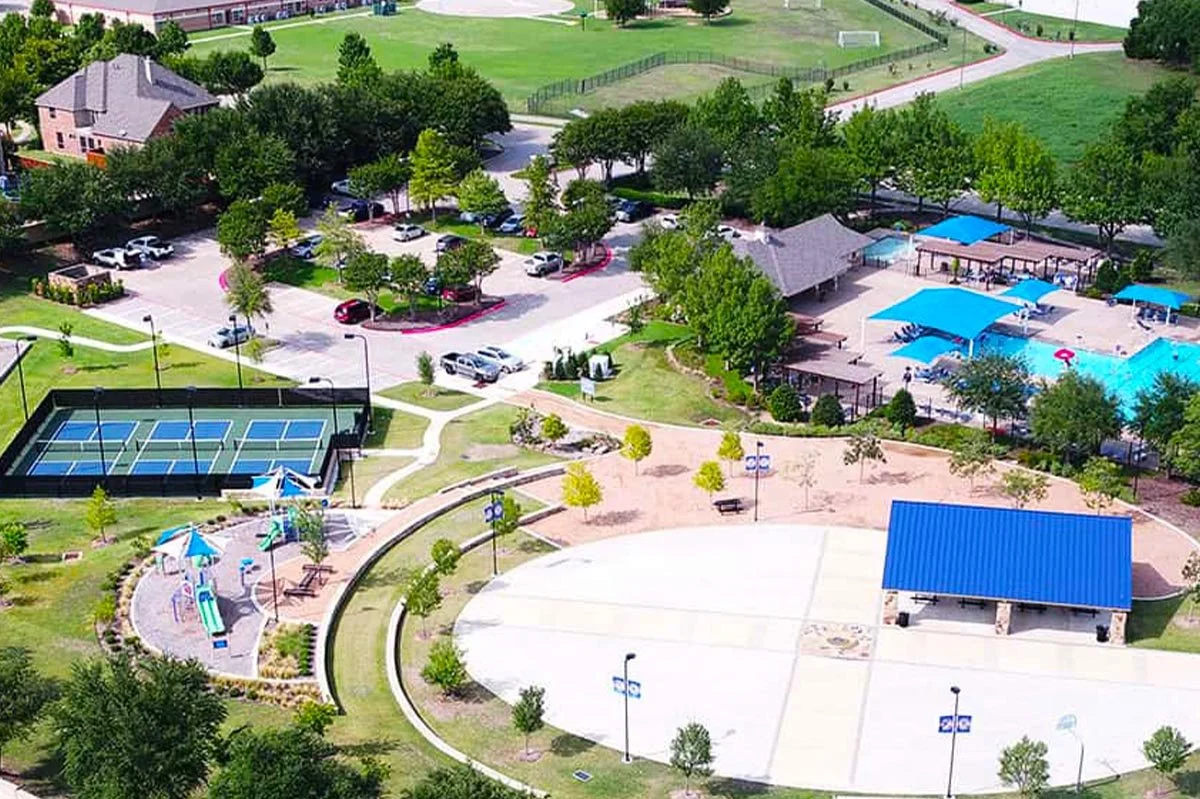 The Stonebridge Ranch Beach Club overlooking Stonebridge Lake in McKinney, Texas. Aerial view of a community park with tennis courts, a swimming pool with blue shade structures, playground, pickleball, and surrounding greenery.