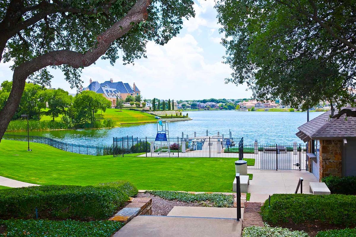 A scenic view of Stonebridge lake with a grassy area, a large tree, a paved walkway, and a fence around a swimming pool near the beach club. In the background, there are houses and trees across the water, under a partly cloudy sky.