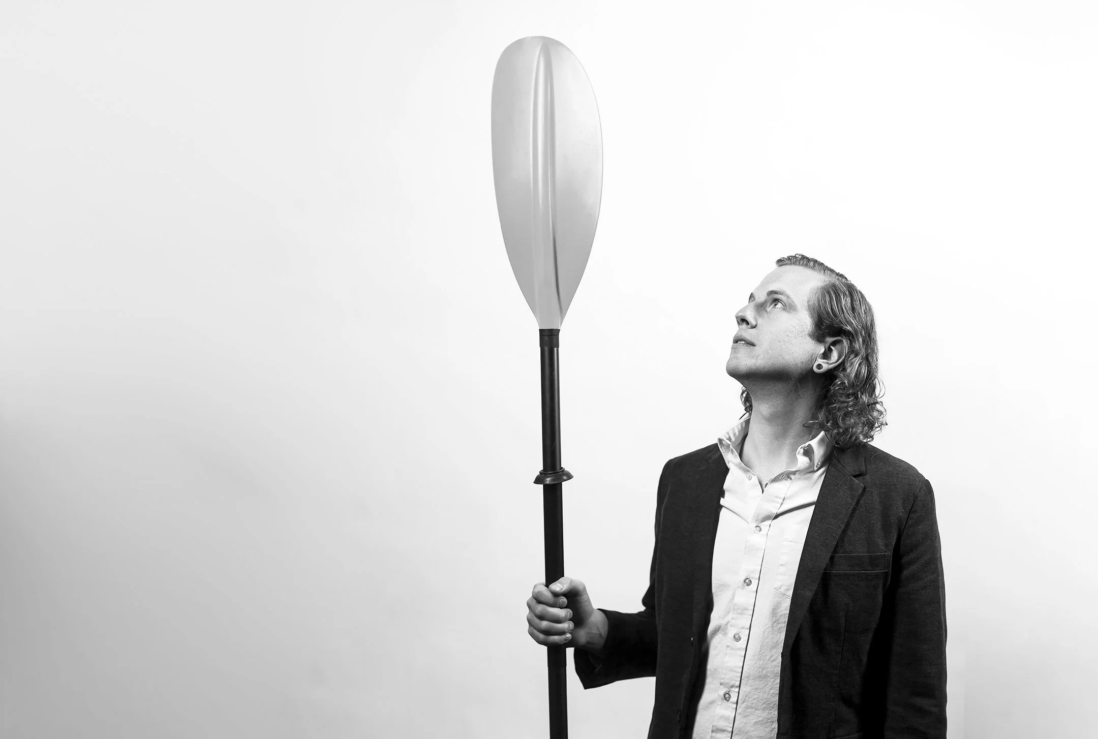 A man with curly hair wearing a suit shirt and jacket, holding a paddle, looking up at a large skateboard leaning against it in a minimalist setting with a plain light background.