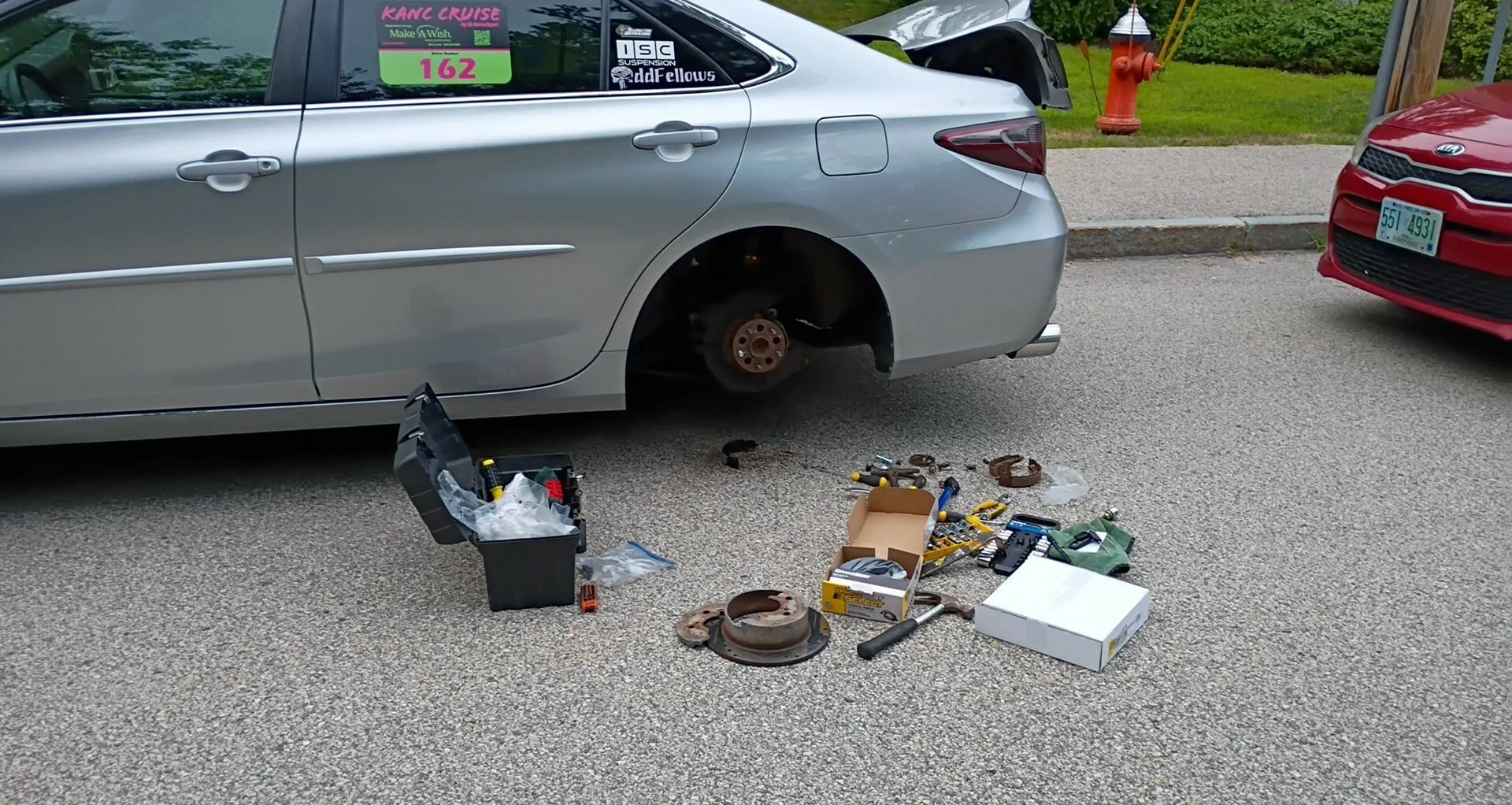 Silver sedan car with its rear wheel removed, parked on a street. Tools and car parts are spread on the ground around it, including a toolbox, brake components, and various hand tools, indicating the car is being repaired or serviced.