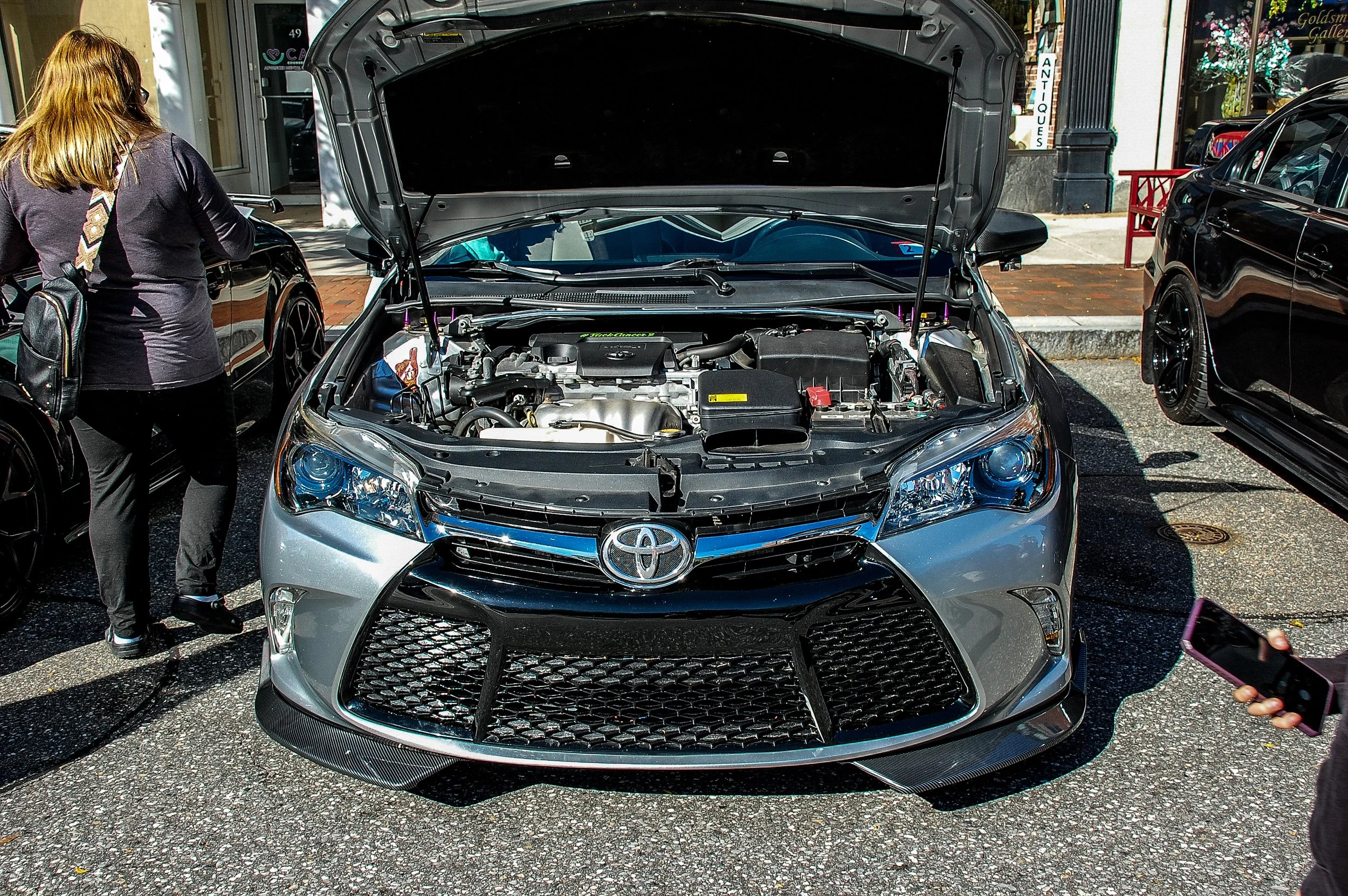 A silver Toyota car with its hood open displaying the engine, parked on a city street. A woman with red hair, wearing a black jacket and pants, is standing nearby.