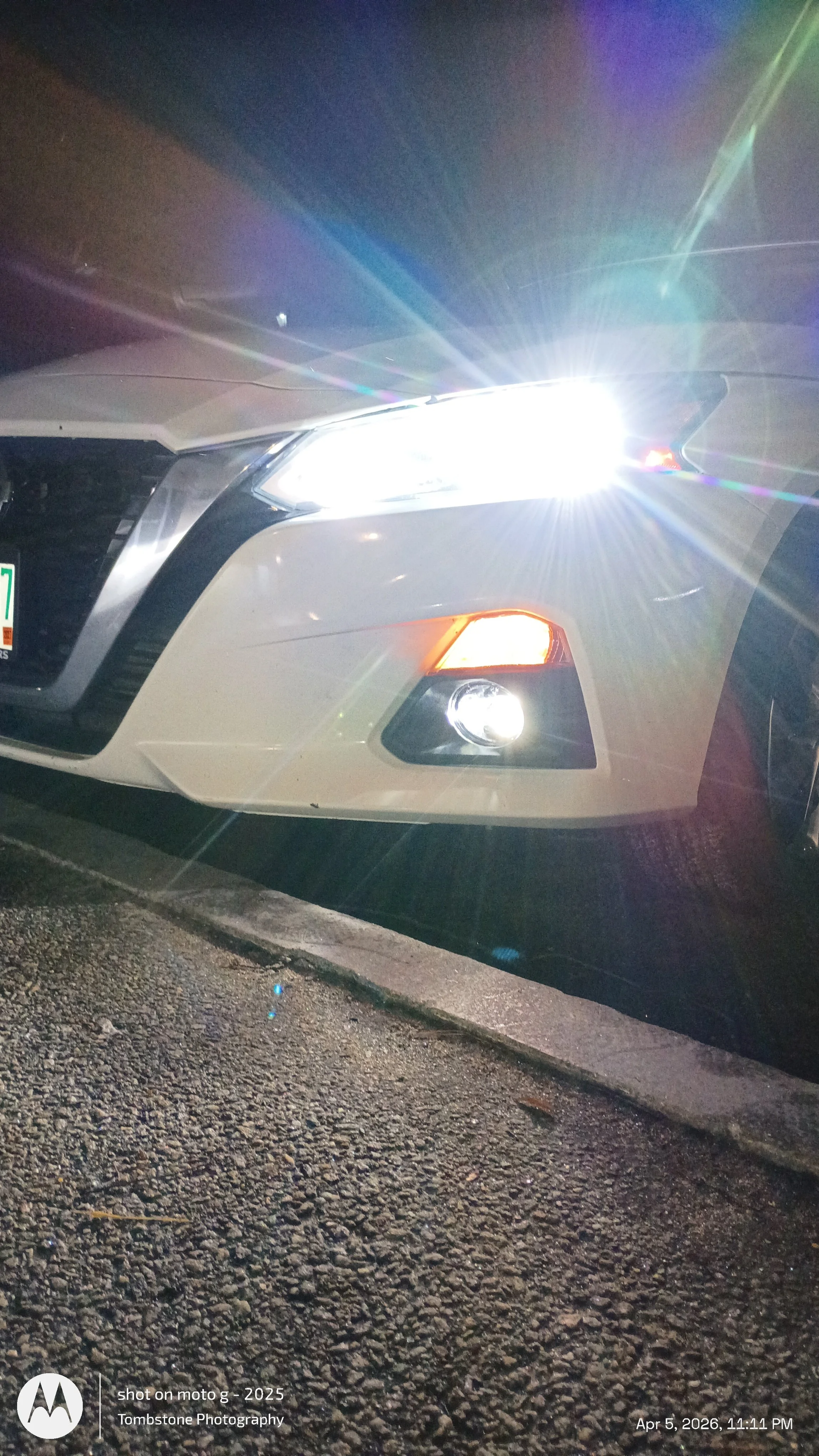 Close-up of a white car's front with bright headlights at night, parked on a gravel surface, with visible lens flares from the headlights.