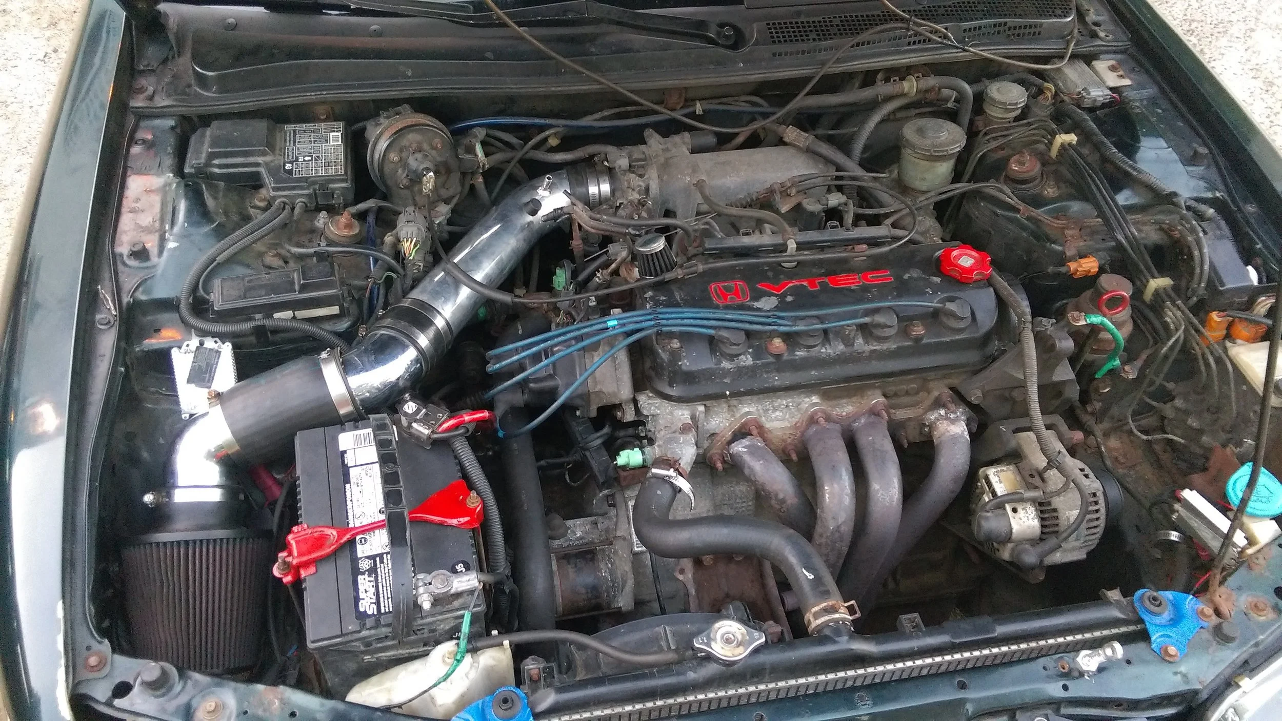 Under the hood of a car showing engine components, including a large chrome intake pipe, a red Honda VTEC valve cover, various hoses, wires, and a black battery with red and black terminals.