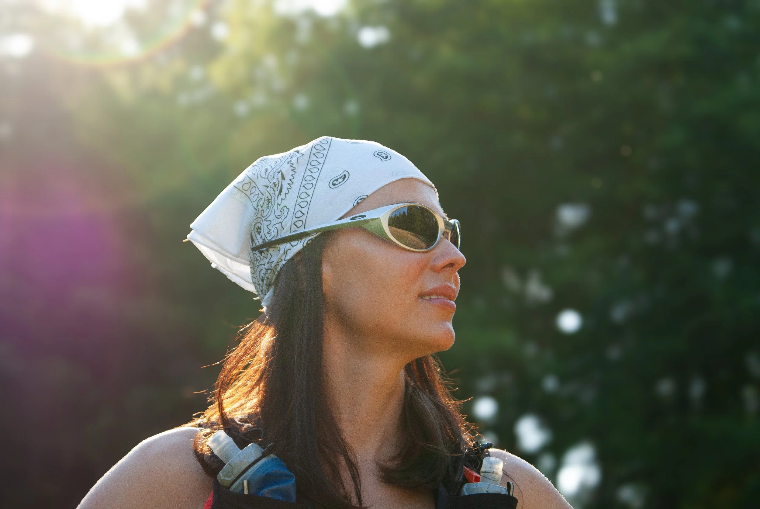 Profile of a woman wearing sunglasses, a white bandana, and a hydration pack outdoors with green blurred trees in the background.
