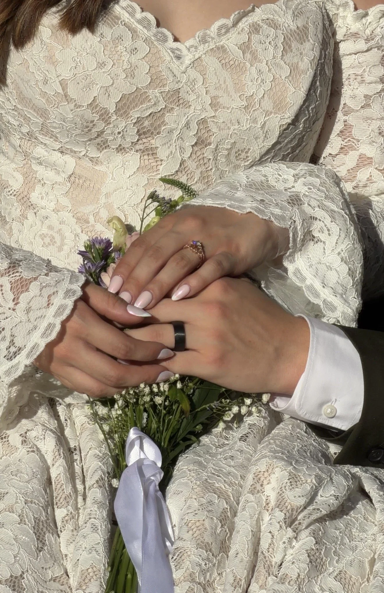 Close-up of a bride and groom's hands, with the bride holding a bouquet of flowers, wearing a cream lace wedding dress, and the groom wearing a white shirt and dark suit.