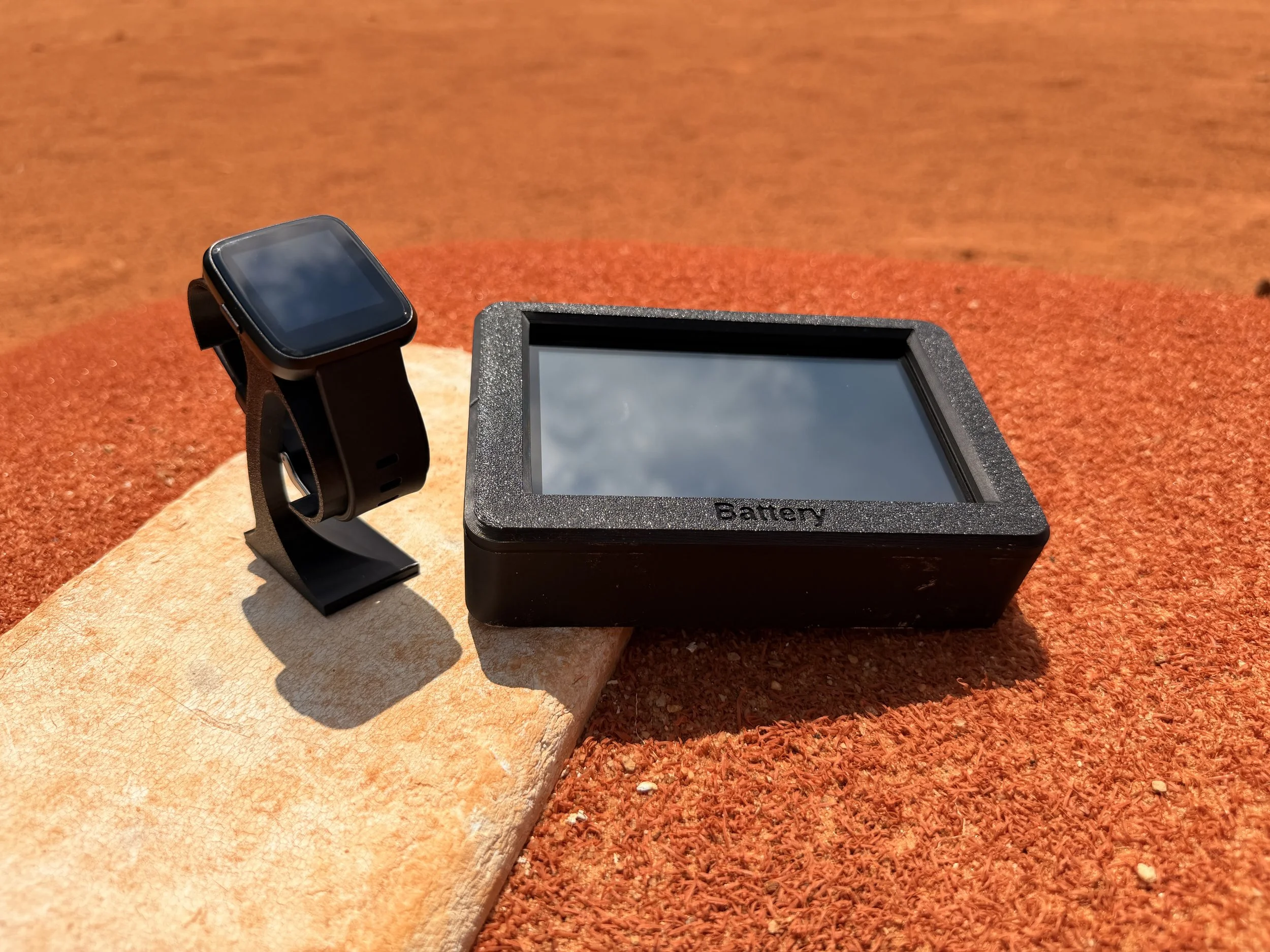 A smartwatch resting on a stand next to a rectangular black device labeled "Battery" on a textured surface, with a reddish-brown background.